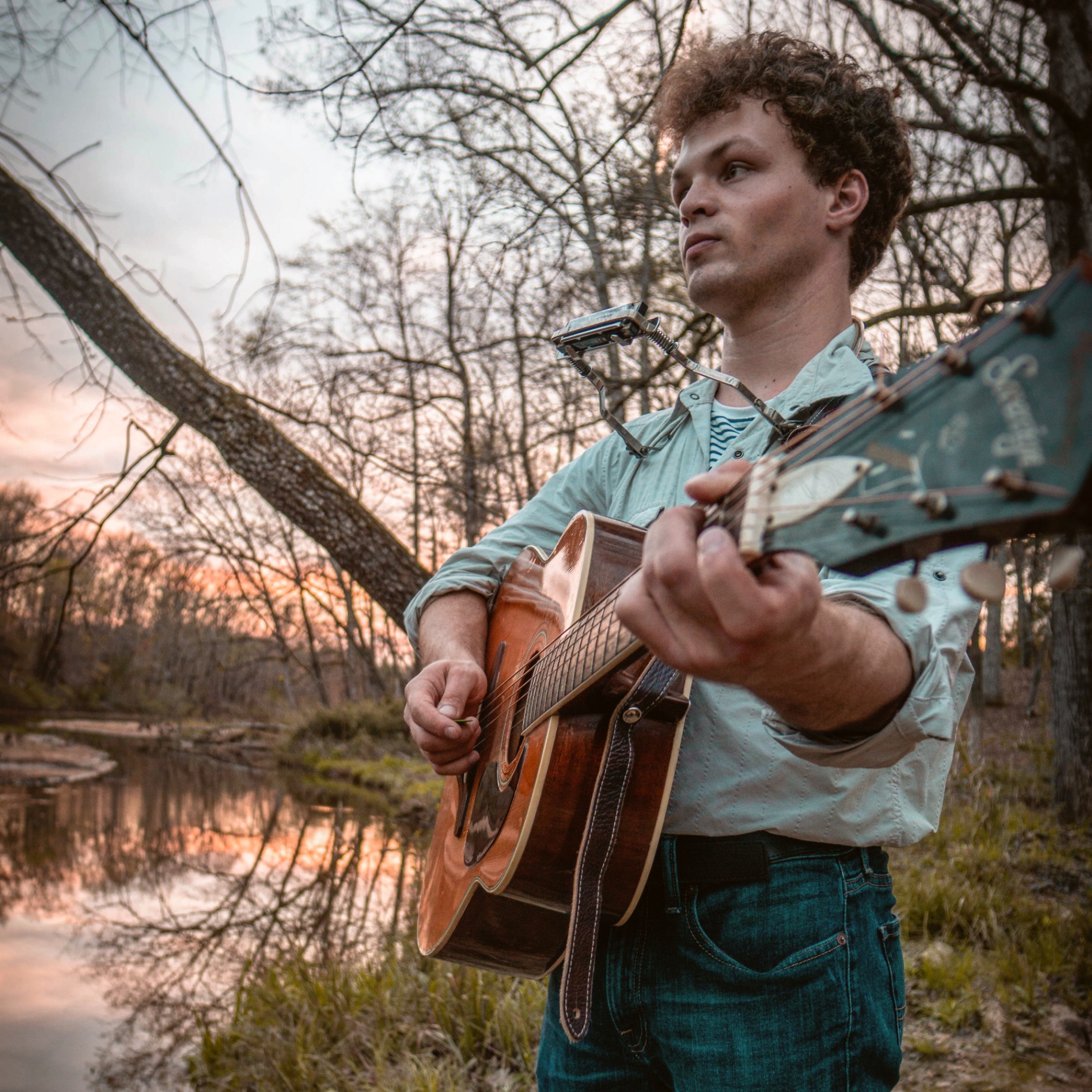 Acoustic guitarist performing outdoors with natural sunset lighting and tree backdrop
