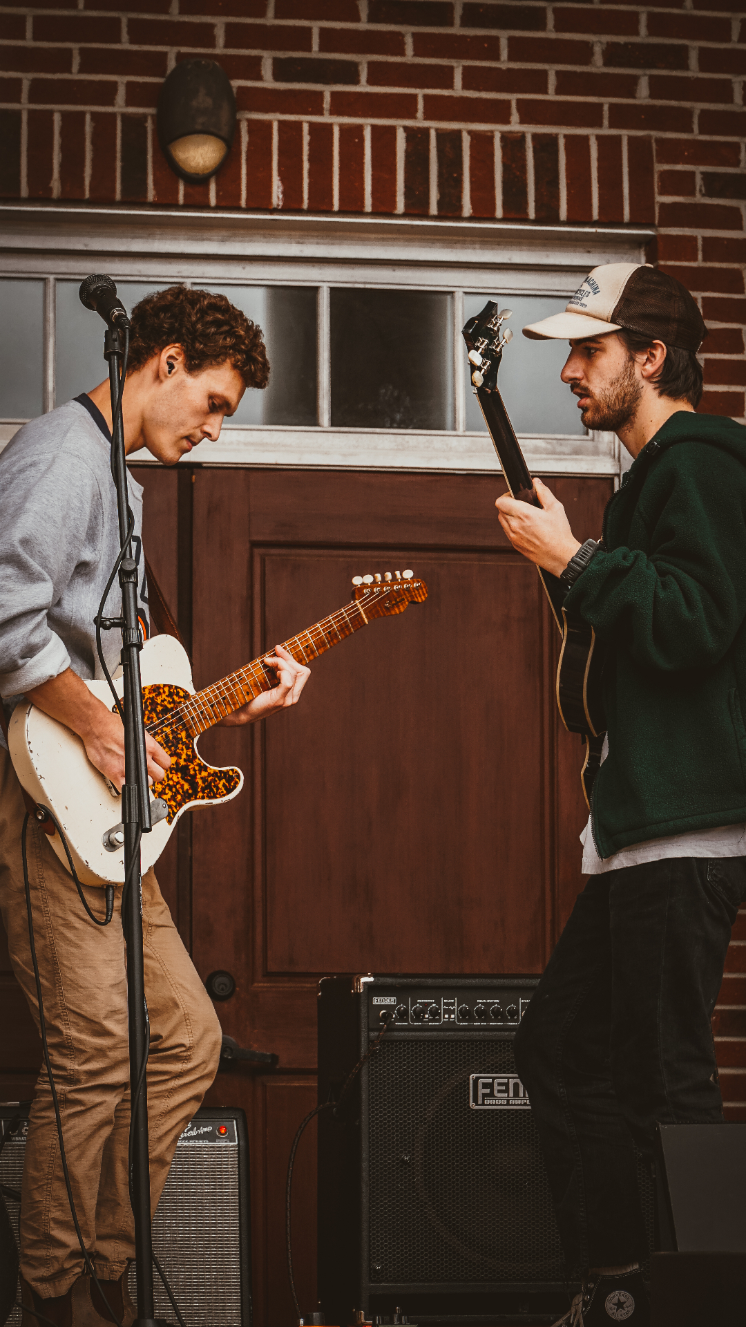 Two guitarists practicing together indoors against brick wall with amplifiers and equipment
