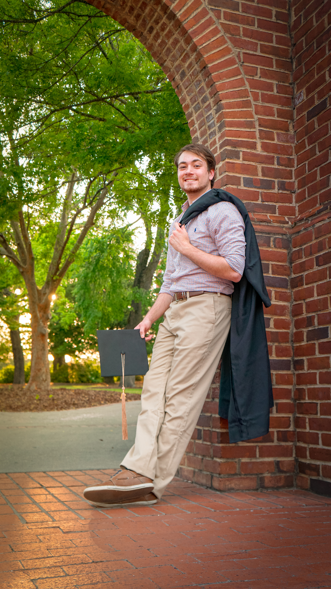 Casual graduation portrait with graduate holding gown under brick archway with greenery