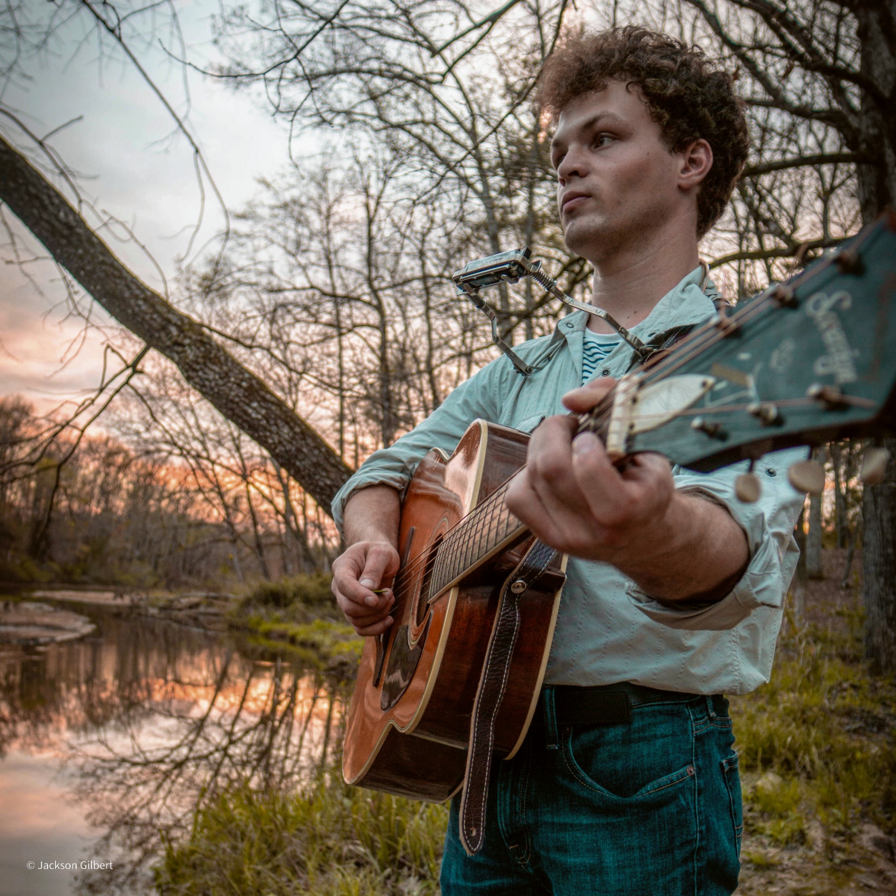Male musician with curly hair playing acoustic guitar outdoors at golden hour with natural tree backdrop