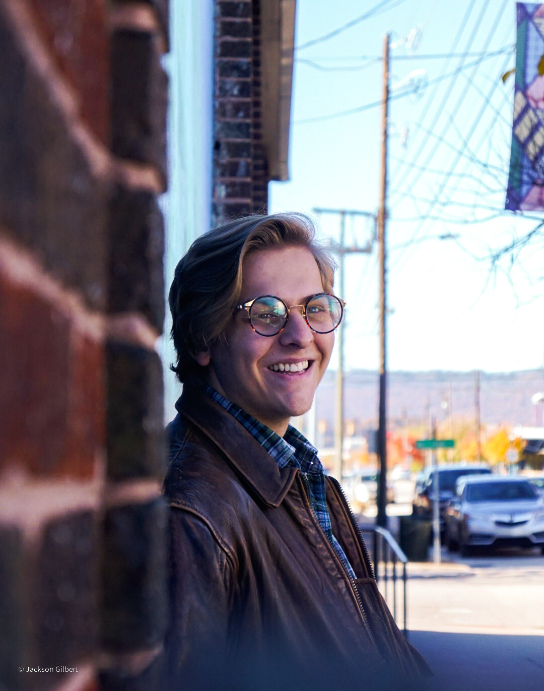 Warm indoor portrait of smiling young man with glasses in leather jacket with natural urban backdrop